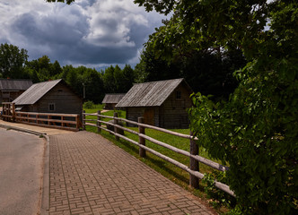 Rural landscape with storm clouds/Road covered with tiles, trees grow. In the foreground there are dirt paths. One-storey wooden buildings are visible. In the background is a cloudy stormy sky. Russia