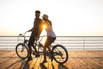 Portrait of a mixed race couple riding on tandem bicycle outdoors near the sea - obrazy, fototapety, plakaty