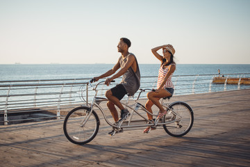 Portrait of a mixed race couple on tandem bicycle outdoors near the sea - obrazy, fototapety, plakaty