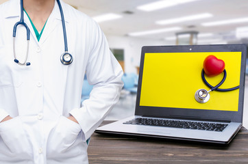 smart doctor with a stethoscope and laptop computer with stethoscope and red heart on yellow screen on wooden desk with blurred hospital background, heart health care and medical technology concept