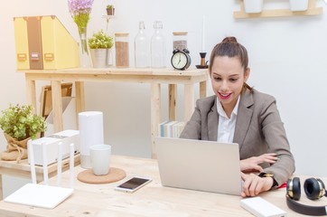 happy asian young business woman beautiful smile working on laptop notebook computer on wood desk at office with alarm clock, headphone, smartphone, wireless router, document folder, soft focus