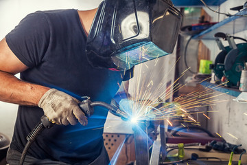 A young male welder in a black T-shirt, black welding mask and construction gloves weld a metal...