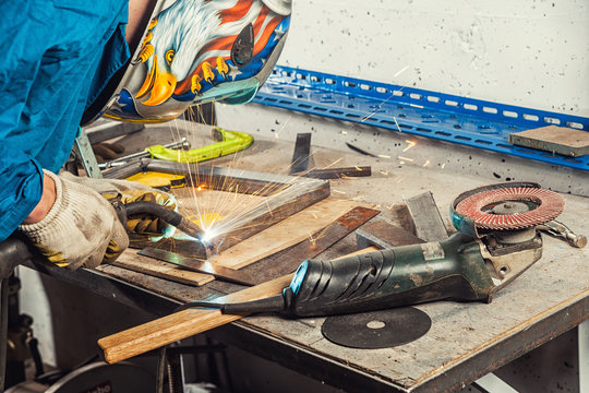 A man welder in a blue  overall, wearing a welding mask with an eagle and construction gloves, weld a metal welding machine in a workshop, sparks fly, instruments and an angle grinder  on the table