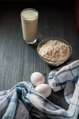 The process of preparation in detail. Cooking, kneading dough. A glass of milk, 2 eggs, a saucer with flour and a blue napkin on a black wooden table