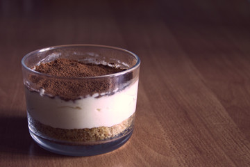 Milk cheesecake in glass, decorated with coffee crumbs, on a wooden table on a blurred background in the early summer morning