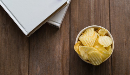 potato chips in a white bowl and book pile on wooden table, view from above