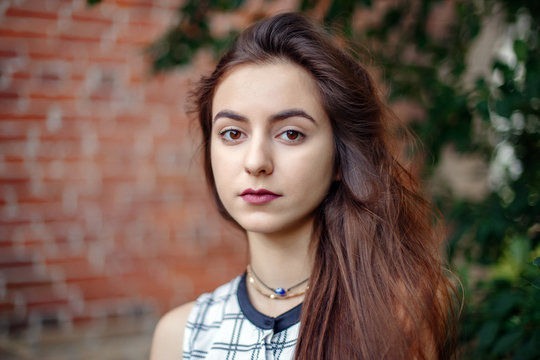 Closeup portrait of beautiful pensive young middle eastern Caucasian jewish woman with long dark hair, black brown eyes, looking in camera. Girl in white plaid shirt against brick wall. Natural beauty