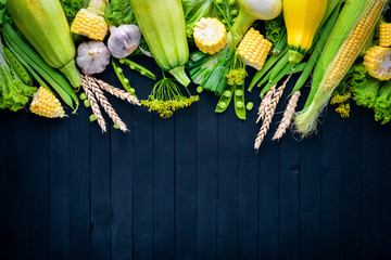 A large selection of raw fresh vegetables. Corn, zucchini, lettuce, greens. On a black wooden background. Top view. Free space for your text.
