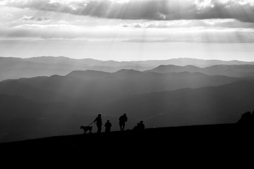 Fototapeta premium Some people and a dog on top of a mountain, with other mountains and hills in the background, and sunrays coming out of the clouds