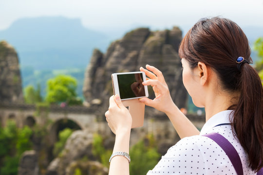 Taking Pictures of Tourist Attraction / Young asian female tourist take photographs with mobile phone of famous Bastei rock formation and bridge in Saxon Switzerland National Park, Germany