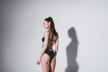 Beautiful slim brunette girl in black swimsuit posing in studio on grey