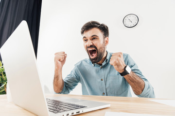 portrait of excited businessman screaming and looking at laptop screen at workplace in office