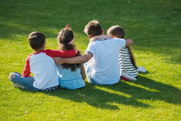 Back view of cute kids sitting embracing on green grass in park