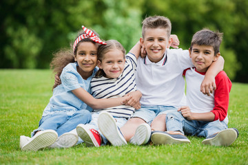 Fototapeta premium Happy multiethnic kids sitting embracing and smiling at camera in park