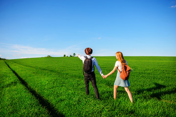 With a backpack, a man in a hat and a woman with long hair go along the path. A couple walks along the meadow