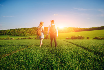 With a backpack, a man in a hat and a woman with long hair go along the path. A couple walks along the meadow