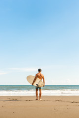 Handsome fit man stand with long surf surfing board wait for wave on surf spot at sea ocean beach. View from back. White blank surfboard. Concept of sport, freedom, new modern life, generation Y.