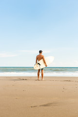 Fototapeta na wymiar Handsome man stand with long surf surfing board wait on surf spot at sea ocean beach. View from back. White blank surfboard. Concept of power, freedom, new modern life, generation Y.