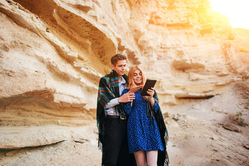 Beautiful woman and handsome man wrapped in a blanket. They are smiling and looking at the screen of a tablet on the background of a sand quarry