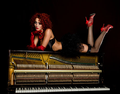 Sexy girl looks like a doll with curly redhead lies on a piano, on a black background. old fashion style