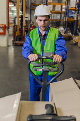 Man with stubble wearing uniform and hardhat steering pallet truck carrying boxes in warehouse. 