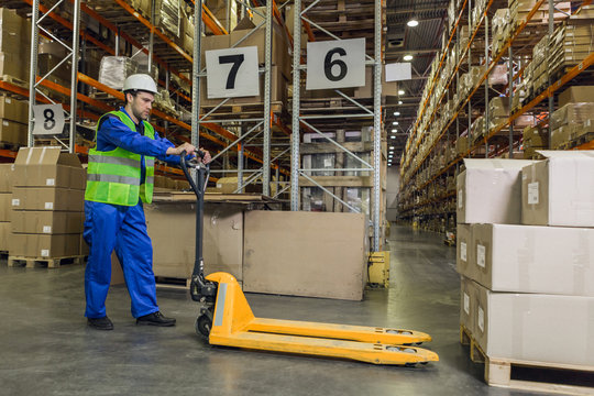 Male worker wearing blue uniform jacket and hardhat driving pallet truck towards pallets with boxes.