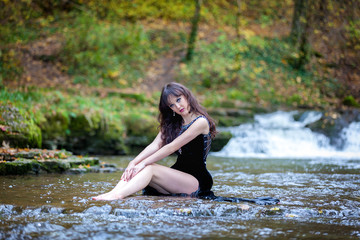 Cute brunette sitting in river raising legs and holding black dress