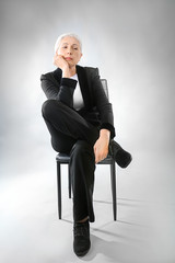 Beautiful elegant elderly woman sitting on chair against gray background