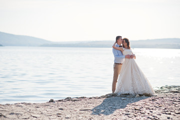 Amazing young couple holding hands on the lake shore on their sunny wedding day.