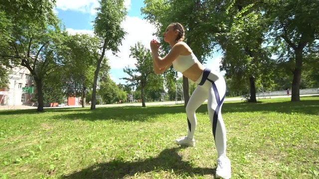Female fitness trainer doing squat exercises outdoors in park in sunny summer morning