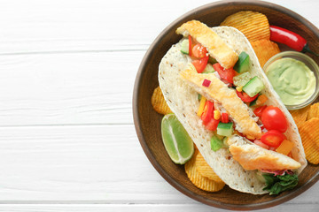 Tasty fish taco on plate with sauce and chips on white wooden background