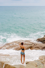 Pretty fit girl in jeans short shorts, red headband and black bra stand on big stone on the beach during sea ocean storm. View from back. Concept of danger, sad, bad emotions.