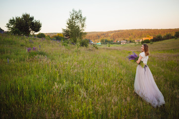 Pretty lady in white dress walks with violet bouquet on green field