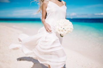 Beautiful blond bride in white wedding dress with white wedding bouquet stand on sea shore. Tropical turquois sea on the background. Summer vacation concept.