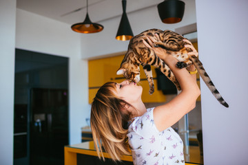 Cheerful woman raises Bengal cat up standing in the kitchen