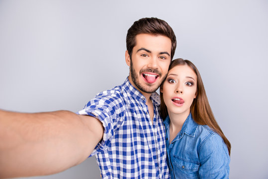 Funky cheerful young couple making selfie photo on guy`s camera. They are in casual outfits, posing and fooling around on pure light background, embracing