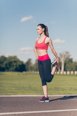Pretty young slim trainer is stretching her legs by doing exercise. She is training outdoors on a summer day, wearing fashionable sport wear, focused
