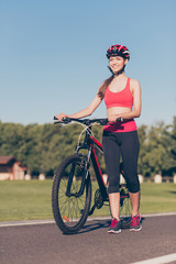 Fototapeta premium Wellbeing and energy concept. Young sporty lady is on a bike outdoors in spring park, wearing helmet and trendy sport outfit, sneakers, smiling