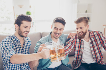 Bachelor men`s life. Close up portrait of three happy men friends, clinking with glasses of beer, smiling, alll are in casual shirts and jeans, sitting on couch at home