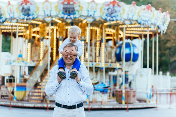 Obraz na płótnie Canvas Father giving son ride on back. Son closes eyes to his Father. They stay over background with carousel in amusement park