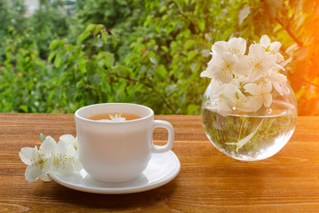 White mug of tea and a vase with jasmine on a wooden table, greens on the background