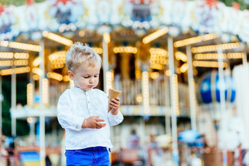 Funny toddler boy in white shirt and blue pants eating an ice cream in front of the carousel © Iryna