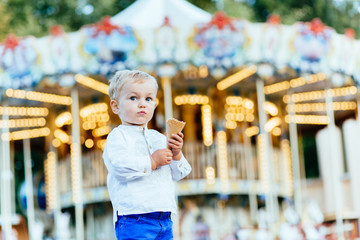 Funny toddler boy in white shirt and blue pants eating an ice cream in front of the carousel