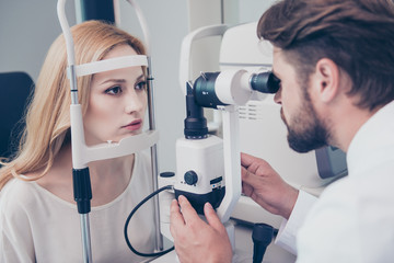 Concentrated brunet bearded optician with non contact tonometer is checking blond`s lady patient intraocular pressure at eye clinic. Health care, medicine, eye sight and technology concept - obrazy, fototapety, plakaty