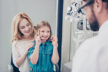 Small cheerful blond girl in jeans outfit is trying on glasses, mom and doc are smiling, she is happy, at the eye lab