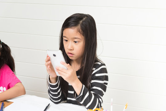 Cute asian girl kid playing smartphone and found something which surprised her while searching on the internet during her study time on light background with friends and copy space