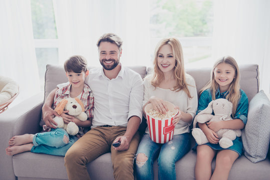 Young beautiful couple of blondie and brunette guy are watching family movie with kids, eating popcorn at light cozy home, smiling, enjoying