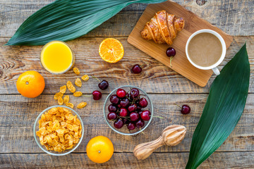 Fototapeta na wymiar Light tasty breakfast. Muesli, oranges, cherry, french croissant and milky coffee on wooden table background top view