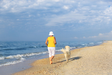 Happy elderly woman running along a beach with her golden retriever at the morning, back view