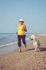 Happy elderly woman running along a beach with her golden retriever at the morning